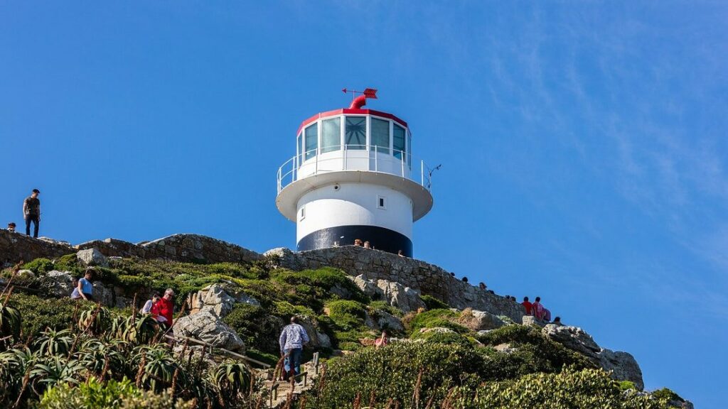 Phare blanc au sommet d'une colline verte, avec quelques visiteurs sur le sentier, sous un ciel bleu