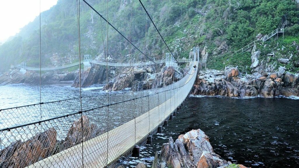 Pont suspendu en bois et grillage au-dessus de l'embouchure d'une rivière, entre des rochers et des pentes boisées