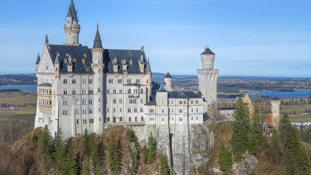Château blanc aux tourelles pointues perché sur un éperon rocheux boisé, avec des lacs et la plaine au fond