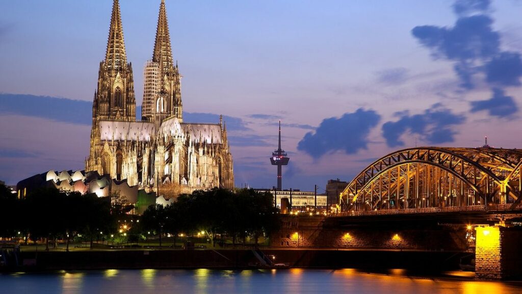 La cathédrale de Cologne et ses deux flèches éclairées près d'un pont ferroviaire illuminé, en fin de journée