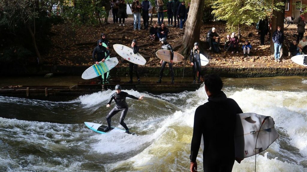 Un surfeur en combinaison sur la vague d'une rivière, avec d'autres planches et des spectateurs sur la berge