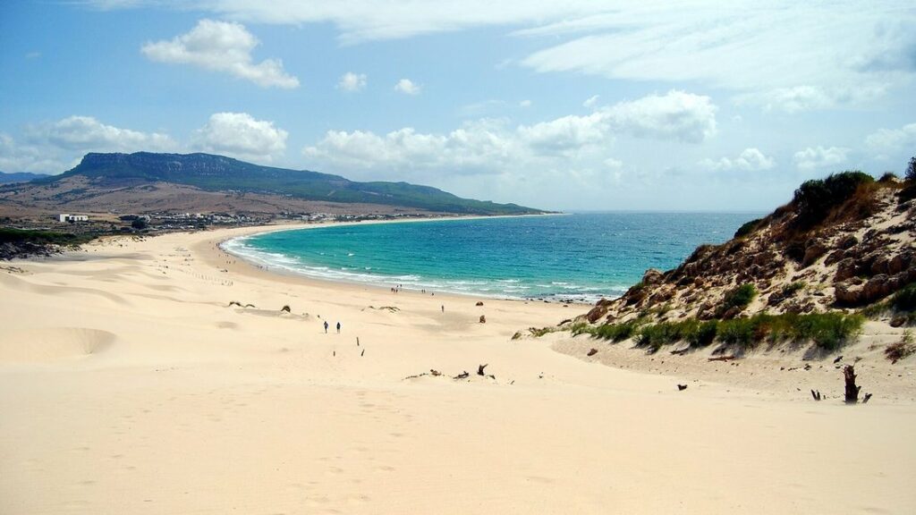 Plage de sable clair et eau turquoise au pied d'une grande dune, avec des montagnes au loin