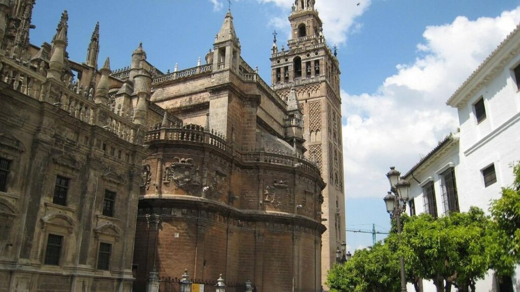 Cathédrale de Séville et sa tour de la Giralda sous un ciel bleu, avec des arbres et des façades blanches au premier plan