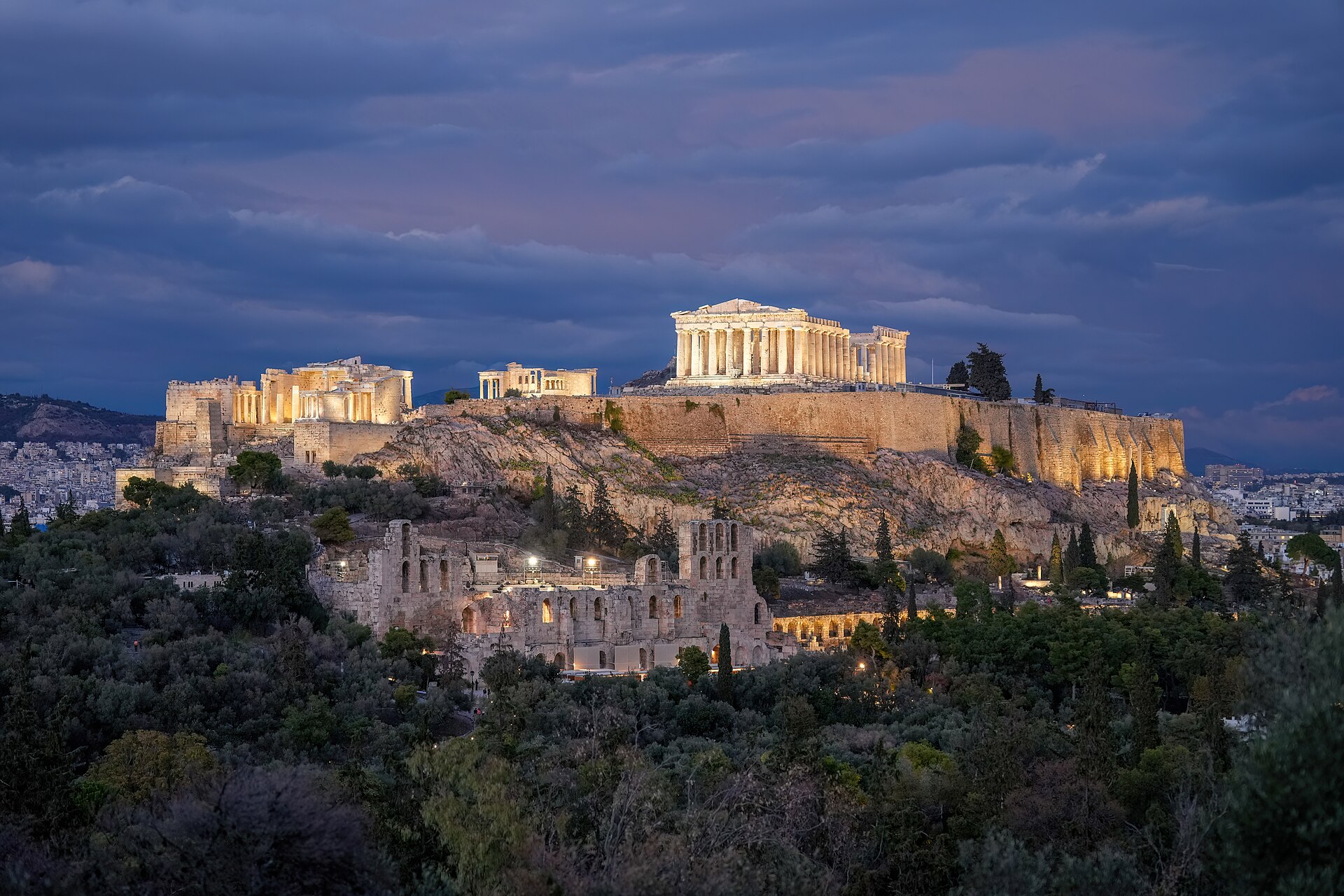 L'Acropole et le Parthénon éclairés au crépuscule sous un ciel sombre, avec l'odéon d'Hérode Atticus au pied de la colline