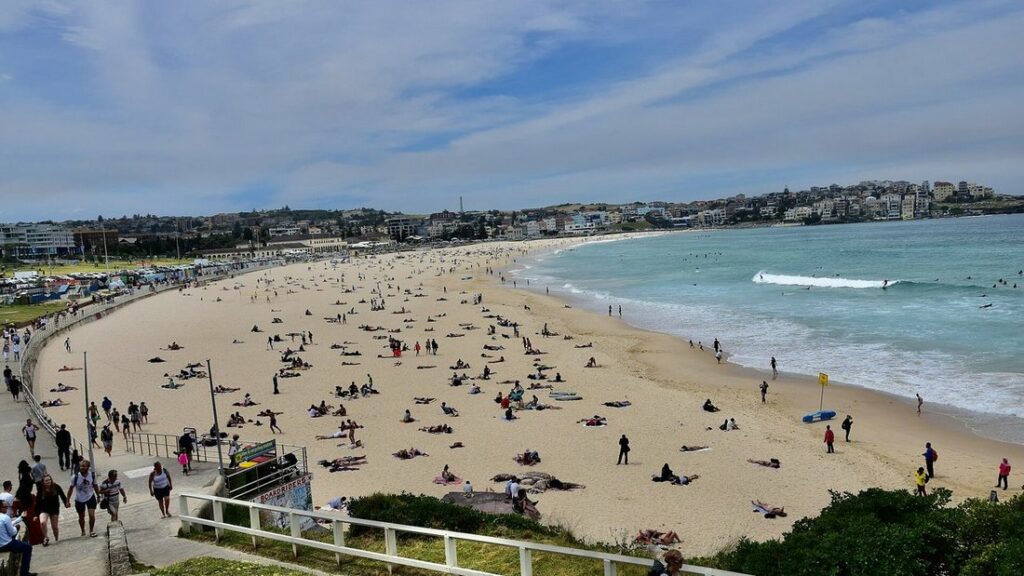 Longue plage de sable clair avec des baigneurs, mer turquoise à droite et immeubles au fond, sous un ciel nuageux