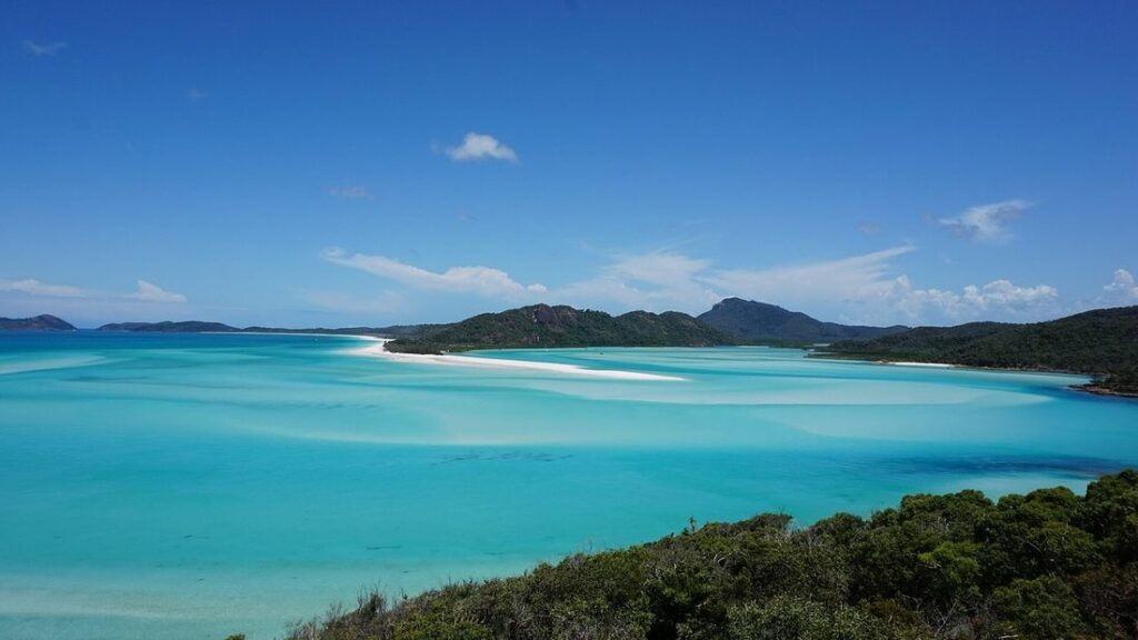 Eau turquoise peu profonde le long d'une plage, avec des collines boisées et des îles au loin sous un ciel bleu