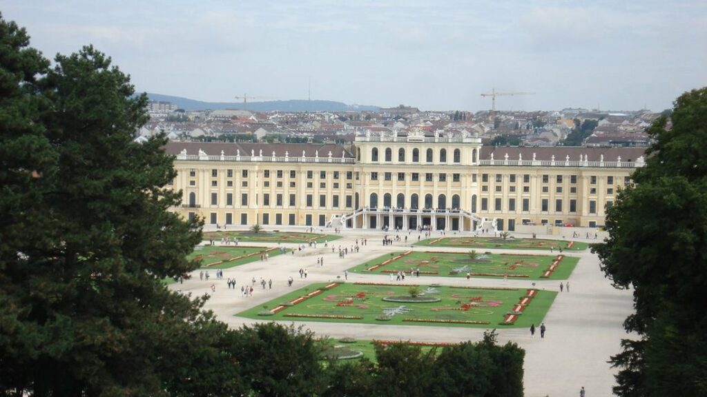Le palais de Schönbrunn et ses parterres vus d'en haut, entre des pins, avec la ville de Vienne derrière