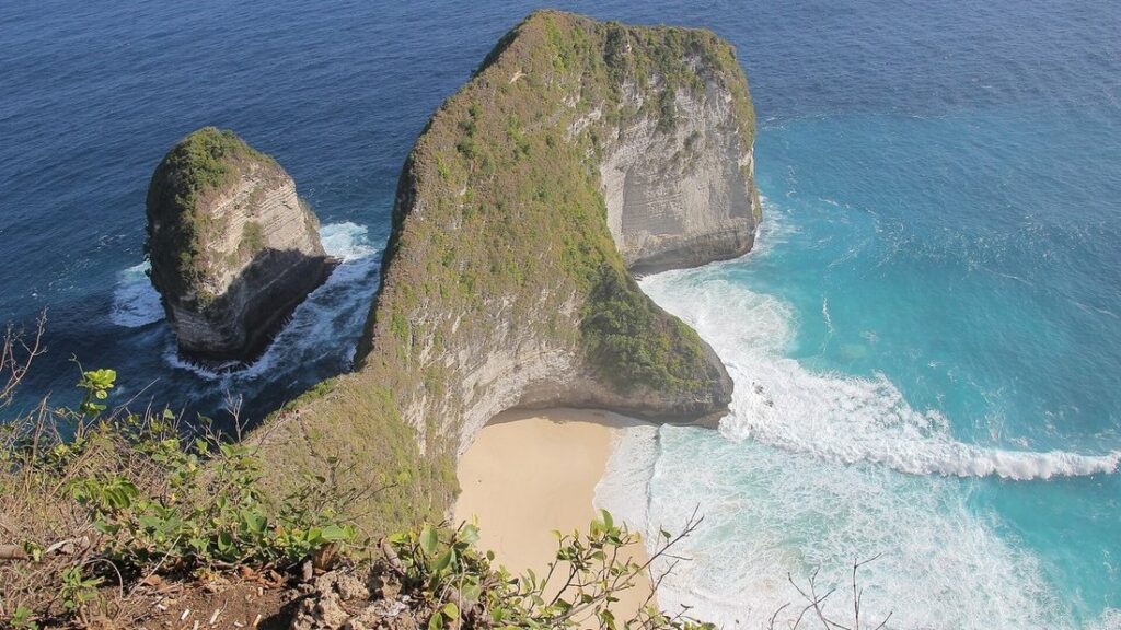 Falaise en forme de promontoire dominant une petite plage de sable et une mer turquoise, avec un îlot rocheux