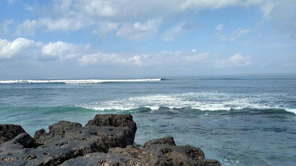 Vagues déferlant devant des rochers sombres, avec de minuscules surfeurs au large, sous un ciel nuageux