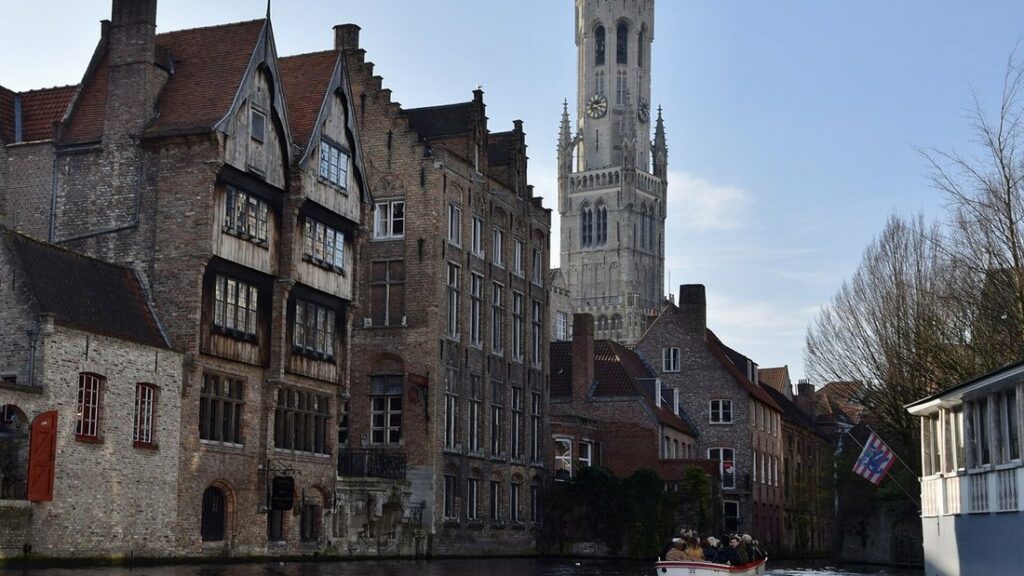Canal bordé de maisons anciennes en brique, avec le beffroi de Bruges qui dépasse des toits
