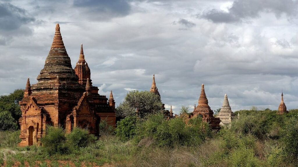 Temple de brique ocre à flèche pointue entouré de végétation, avec d'autres pagodes au loin sous un ciel nuageux