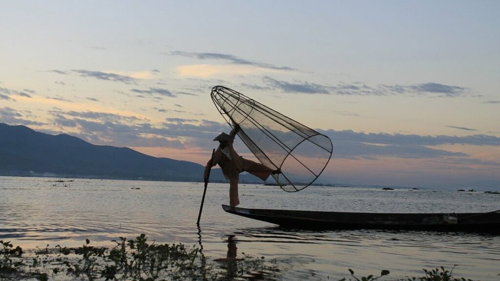 Pêcheur debout sur une jambe à l'arrière de sa barque, avec une nasse conique, sur le lac Inle au crépuscule