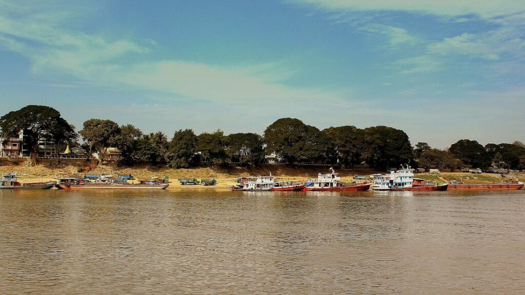 Bateaux amarrés le long de la rive boisée de l'Irrawaddy, sous un ciel bleu, avec l'eau brune du fleuve au premier plan