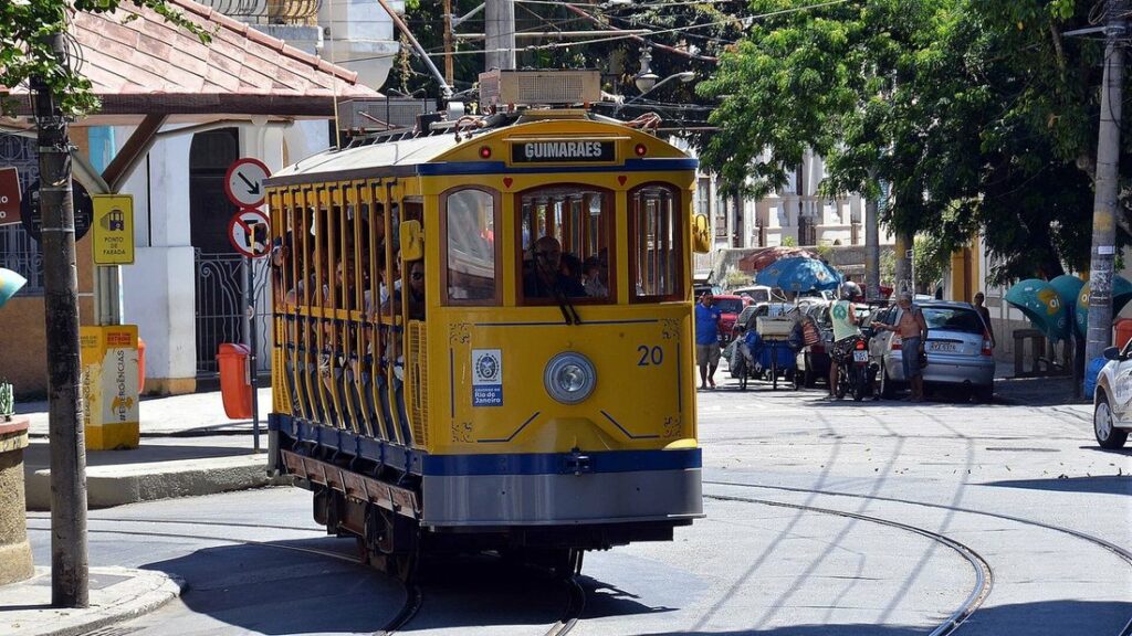 Tramway jaune ouvert de Santa Teresa, passagers debout sur les côtés, dans une rue de quartier ensoleillée