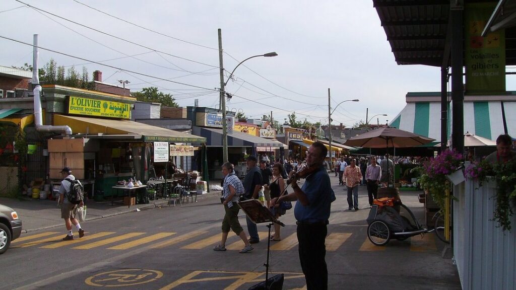 Rue du marché Jean-Talon bordée d'étals sous auvents, avec des passants et un violoniste au premier plan