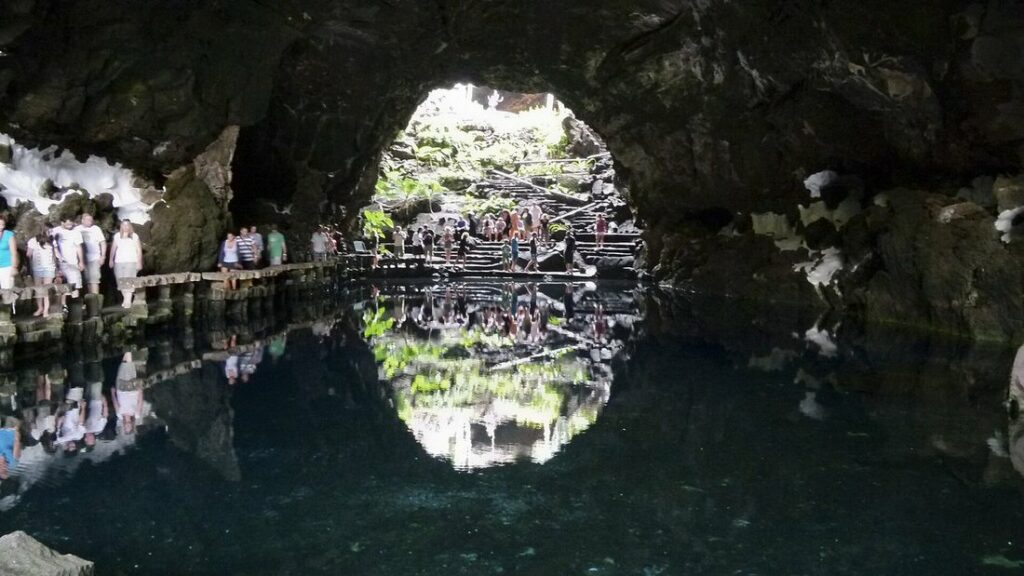 Lac aux eaux sombres dans une grotte volcanique, avec une ouverture dans la voûte et des visiteurs sur un passage