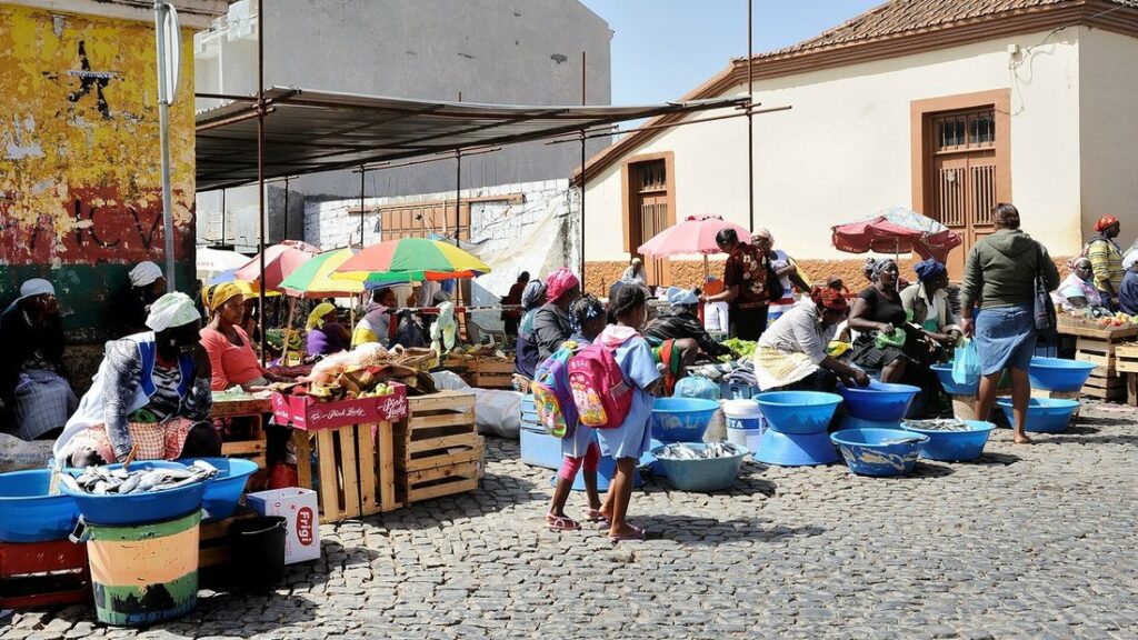 Étals sous parasols colorés sur une place pavée d'un marché de rue, avec des vendeuses et des clients