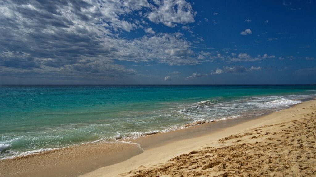 Plage de sable clair léchée par des vagues turquoise, sous un ciel bleu chargé de gros nuages