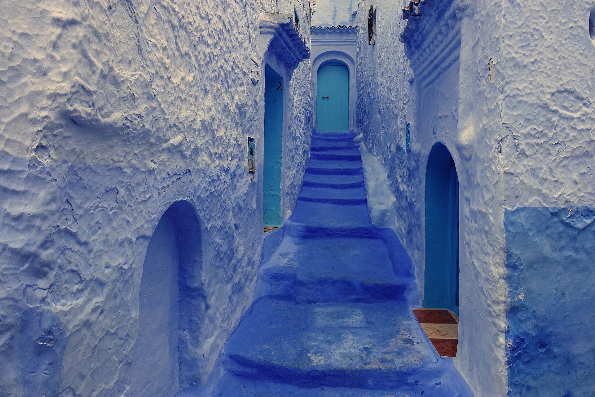 Escalier de ruelle peint en bleu entre deux murs chaulés aux reflets irréguliers, avec des portes turquoise au fond
