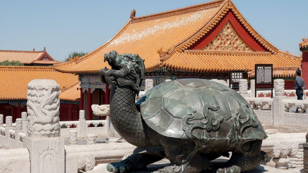 Tortue de bronze à tête de dragon devant un pavillon aux toits de tuiles jaunes et aux avant-toits peints