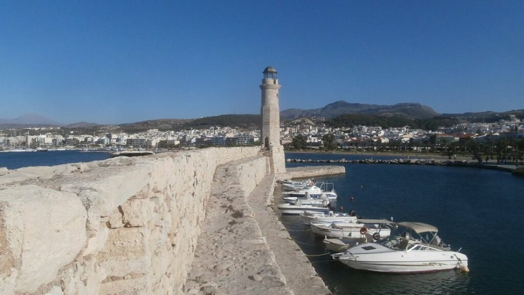 Phare de pierre au bout de la jetée du port vénitien de Réthymnon, avec des bateaux amarrés et la ville au fond