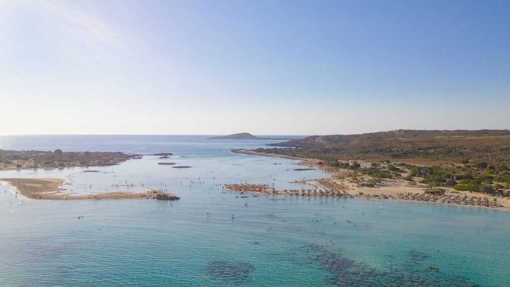 Vue aérienne du lagon d'Elafonissi, eaux claires peu profondes, parasols sur la plage et côte rocheuse au fond