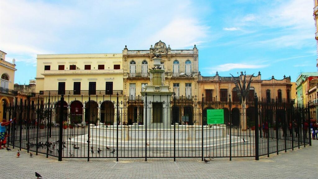 Place bordée de façades coloniales aux couleurs claires, avec une fontaine derrière une grille de fer