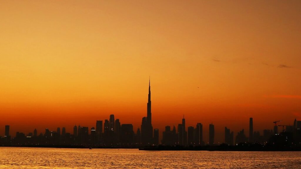 Silhouette de la tour Burj Khalifa dominant les gratte-ciel de Dubai sous un ciel orange, au-dessus de l'eau