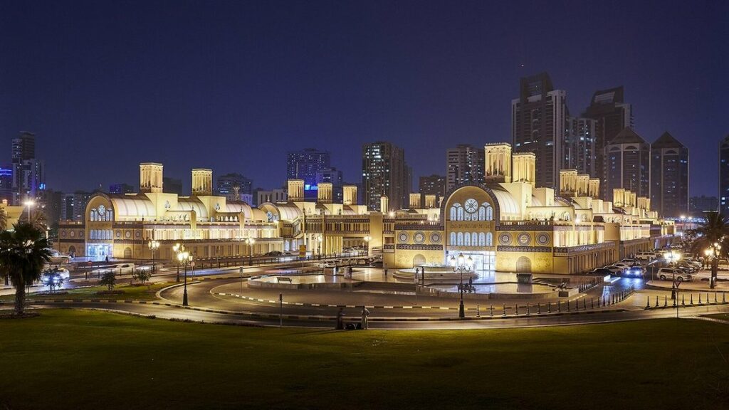 Le Souk al-Markazi de Sharjah illuminé de nuit, bâtiment à tourelles et arcades, avec des tours en arrière-plan