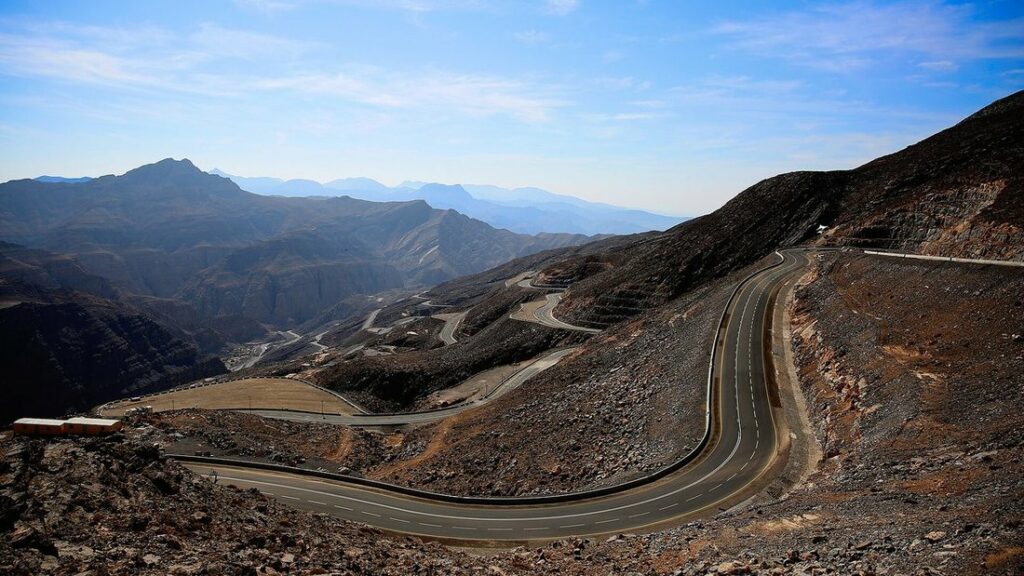 Route sinueuse montant dans les montagnes brunes et rocailleuses du Jebel Jais, sous un ciel bleu