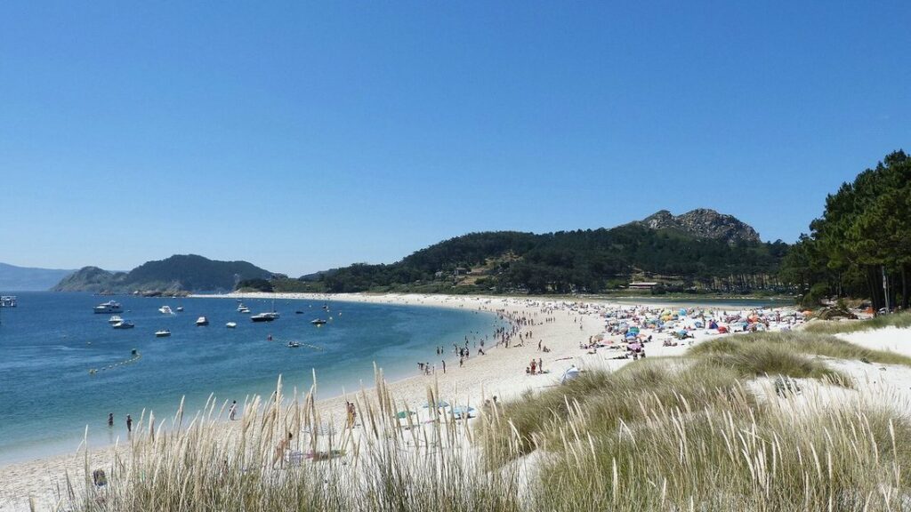 Longue plage de sable clair bordée de dunes et de collines vertes, avec quelques bateaux sur l'eau bleue