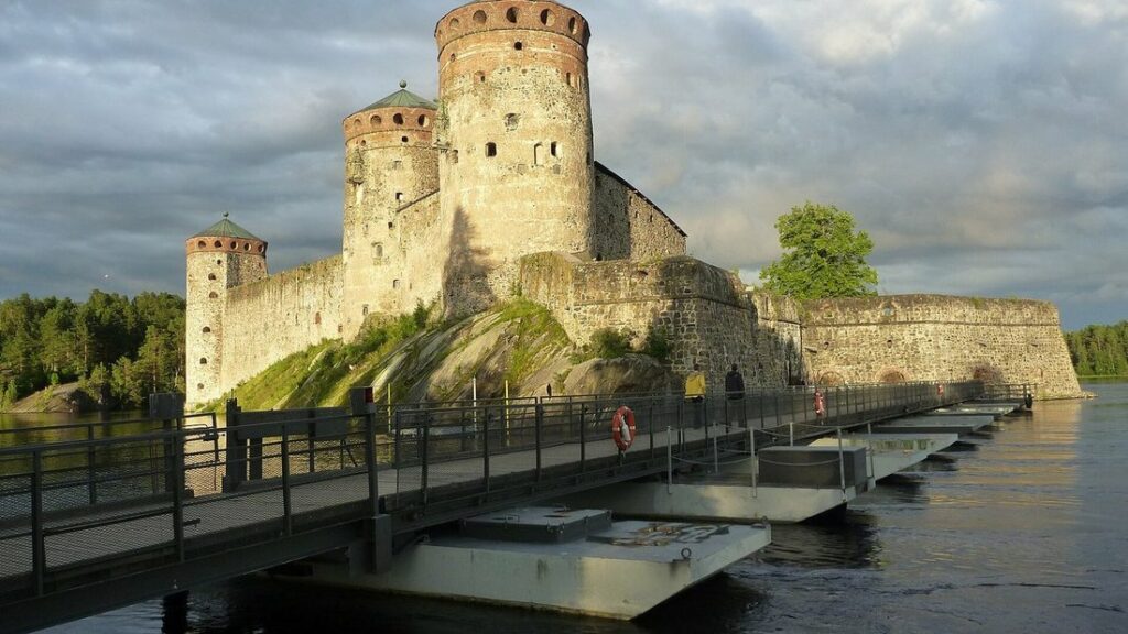 Château d'Olavinlinna en pierre claire avec ses tours rondes, relié à la rive par un pont flottant