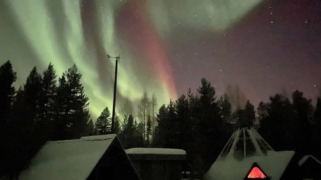 Aurore boréale verte et rouge au-dessus d'une forêt sombre et d'une cabane de bois de type kota