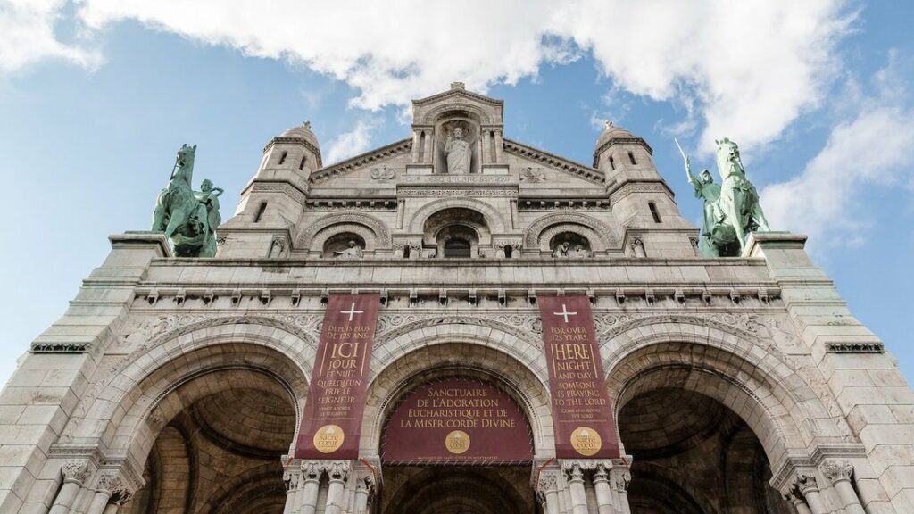 Façade blanche d'une basilique à portails en arcades, vue d'en bas, avec deux statues équestres vertes sous un ciel bleu nuageux