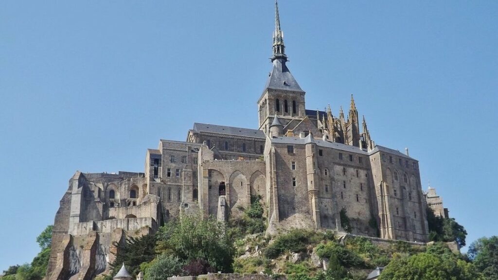 Abbaye de pierre surmontée d'une flèche, perchée sur un rocher couvert d'arbres, sous un ciel bleu