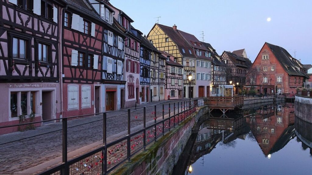 Maisons à colombages colorées alignées le long d'un canal, avec leur reflet dans l'eau et une rambarde au premier plan