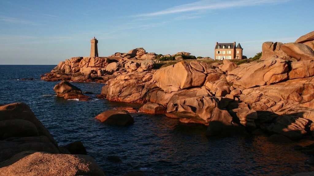 Rochers de granit rose éclairés par une lumière rasante, avec un phare et une maison sur la pointe, au bord de la mer