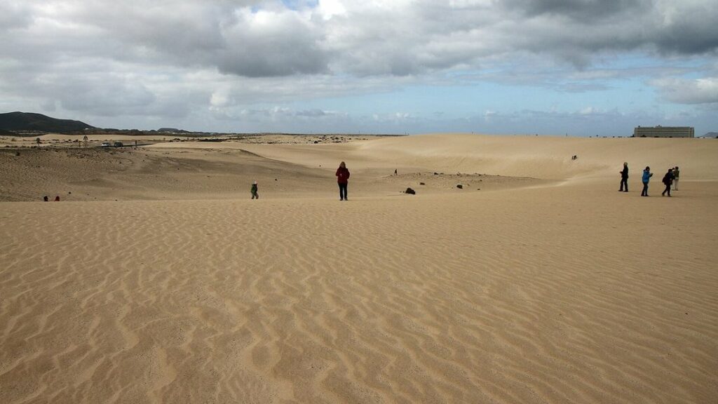 Vaste étendue de sable doré ridé par le vent, avec quelques promeneurs au loin sous un ciel nuageux