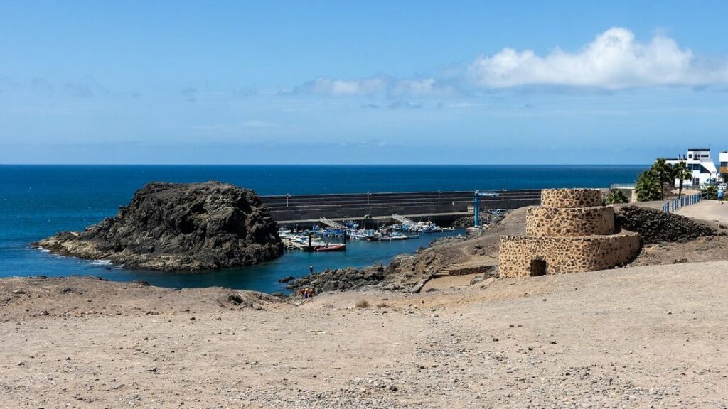 Rocher volcanique noir devant un petit port abrité par une digue, avec une construction de pierre ronde à droite
