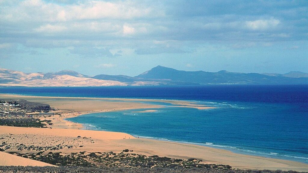 Longue plage de sable clair et eau bleue vue de haut, avec des reliefs ocre au fond