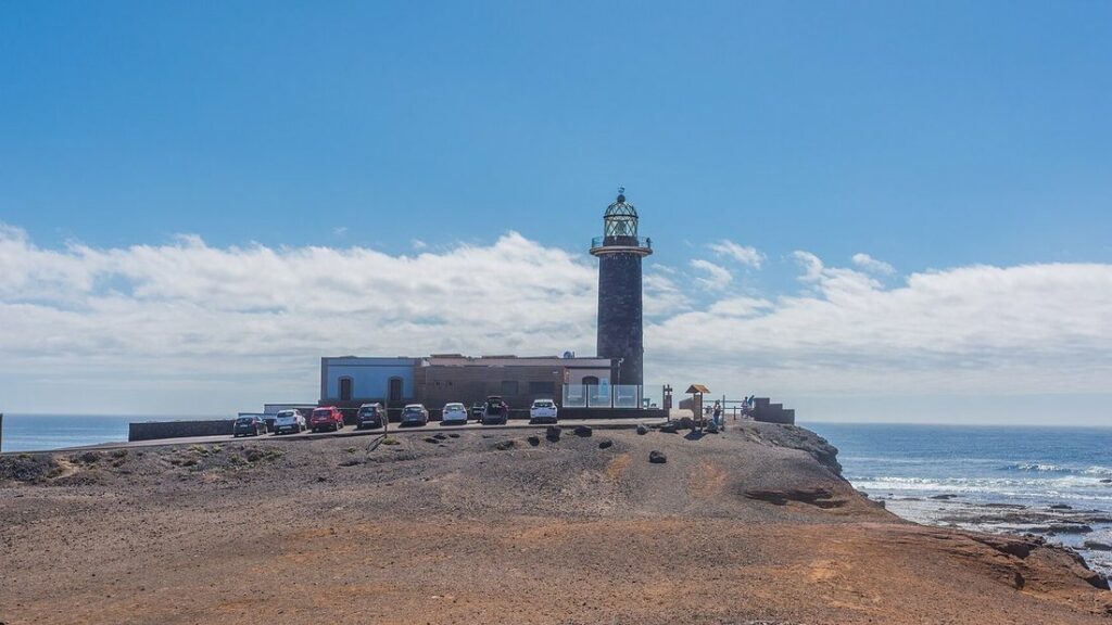 Phare sombre et bâtiments blancs sur une pointe rocheuse au bord de l'océan, sous un ciel bleu