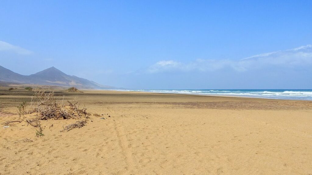 Vaste plage de sable doré ouverte sur la mer, avec un relief sombre au fond sous un ciel bleu