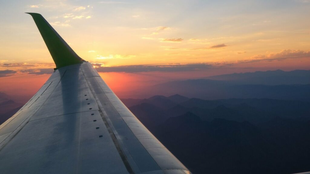Aile d'avion à l'ailette verte au coucher du soleil, au-dessus d'un relief de montagnes dans la brume