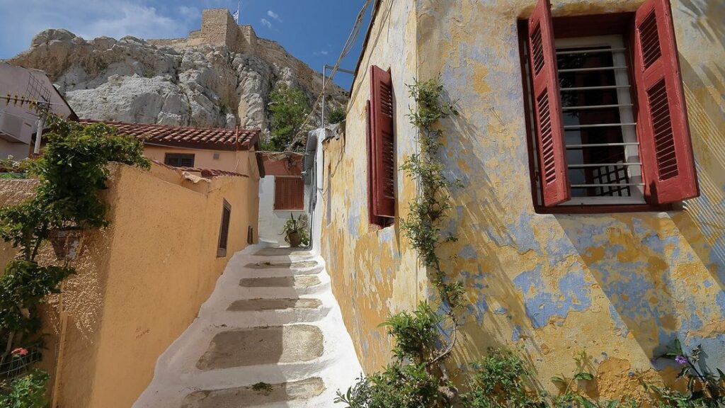 Ruelle d'Anafiotika avec escalier blanc, murs ocre aux volets rouges et rocher de l'Acropole en arrière-plan