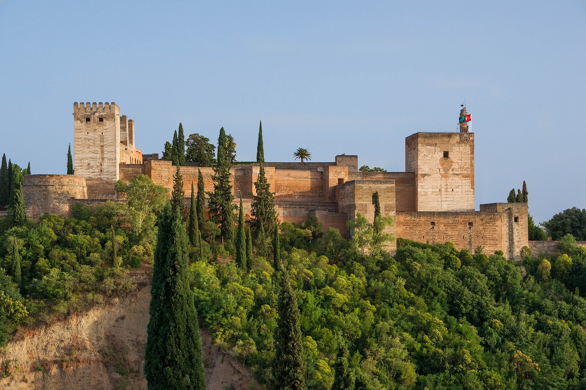Tours et remparts de pierre rouge de l'Alhambra au sommet d'une colline plantée de cyprès, sous un ciel bleu