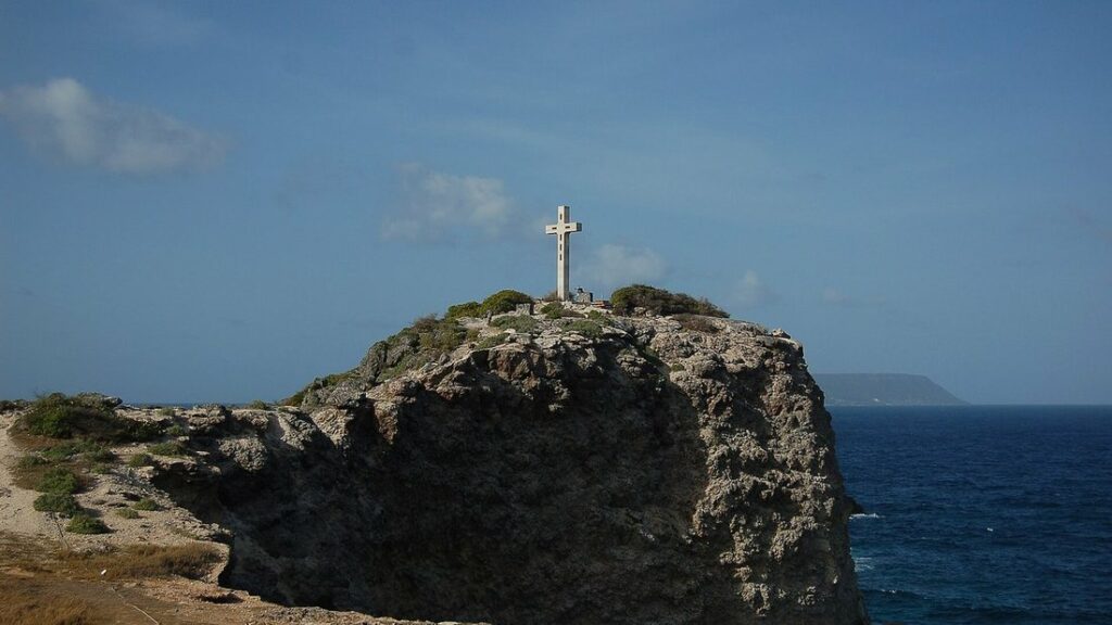 Rocher sombre surmonté d'une croix blanche sur la pointe, face à l'océan bleu et à un ciel voilé