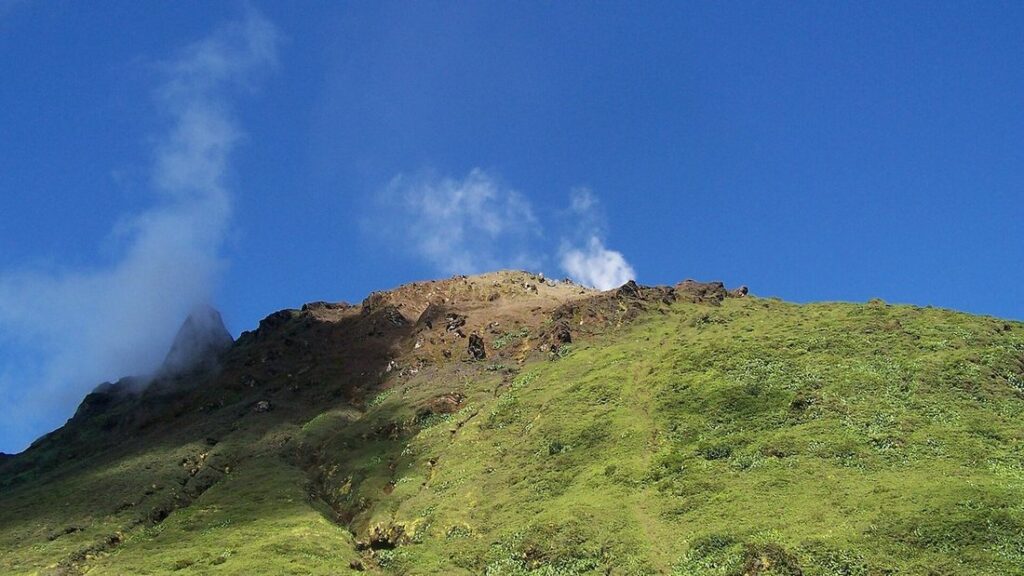 Pente couverte d'herbe verte et sommet brun du volcan de la Soufrière, avec un panache de vapeur sous un ciel bleu