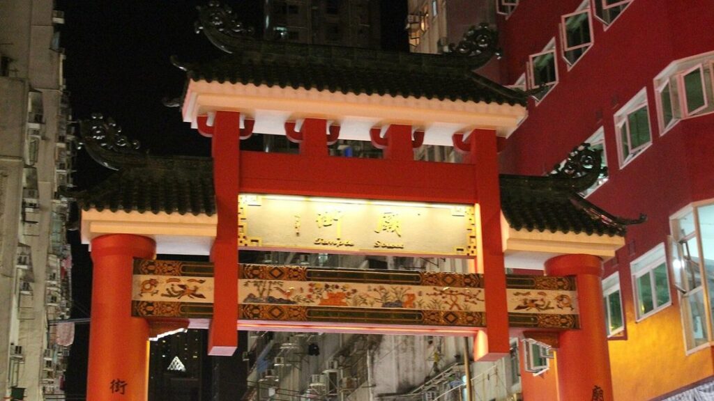 Portique rouge et blanc à toit de tuiles, éclairé de nuit, à l'entrée de Temple Street entre des immeubles