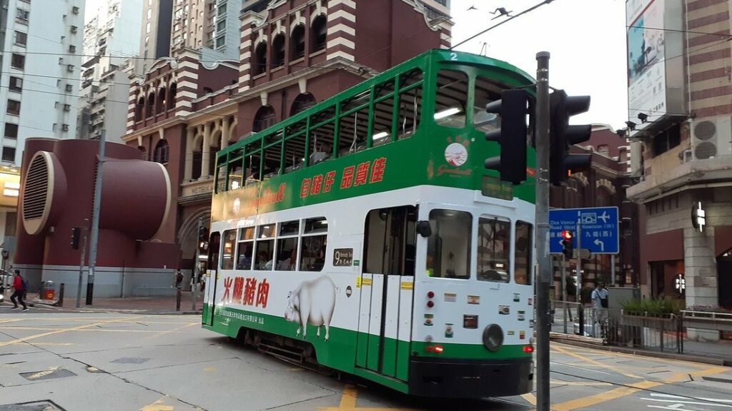 Un tram à impériale vert et blanc tourne à un carrefour, devant un bâtiment de brique rouge et blanche