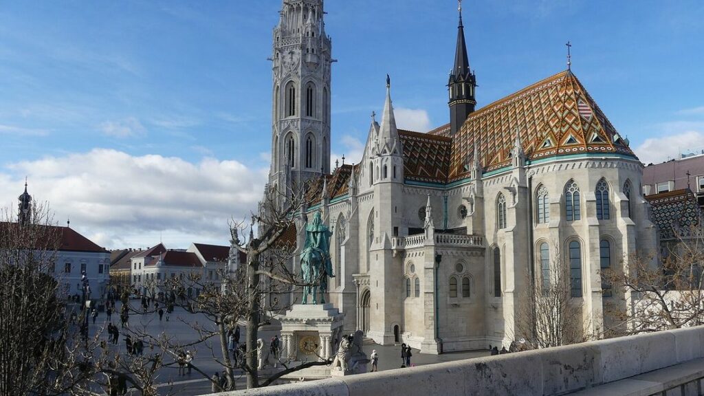 Église Matthias aux murs de pierre claire et au toit de tuiles colorées, avec une statue équestre verte au premier plan